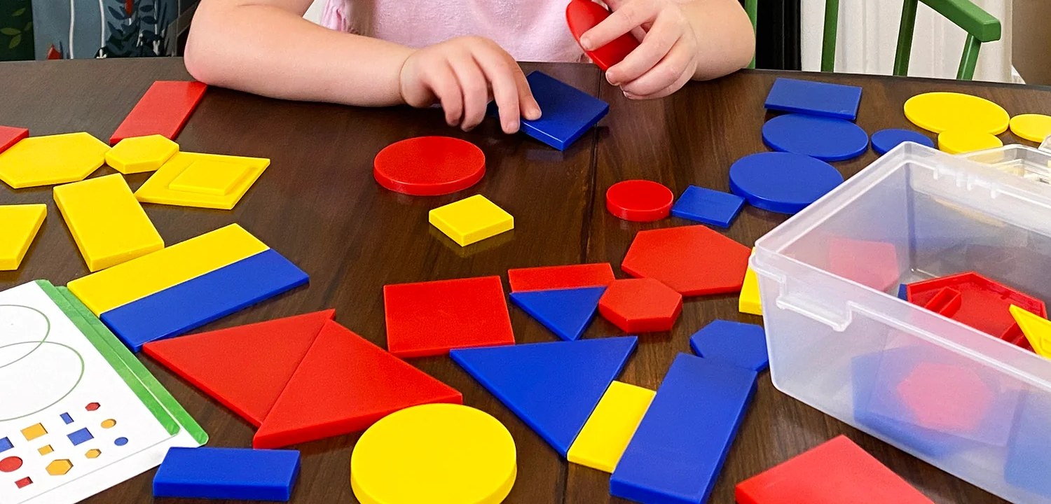Child's hands sorting colorful red, blue, and yellow attribute blocks on a wooden table