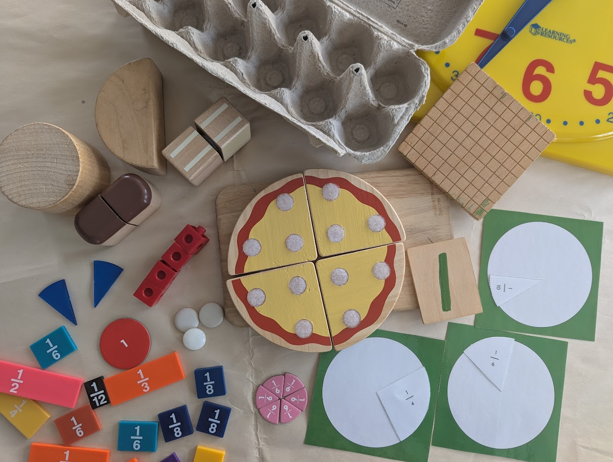 A collection of fraction materials spread out: fraction circles, a toy pizza with wedges, number cards, eggs, wooden shapes, and grid paper