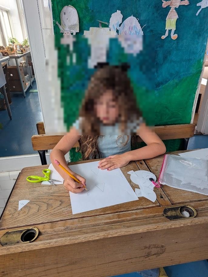 A child working at a wooden desk, cutting paper shapes with scissors, with children's artwork visible on the wall behind
