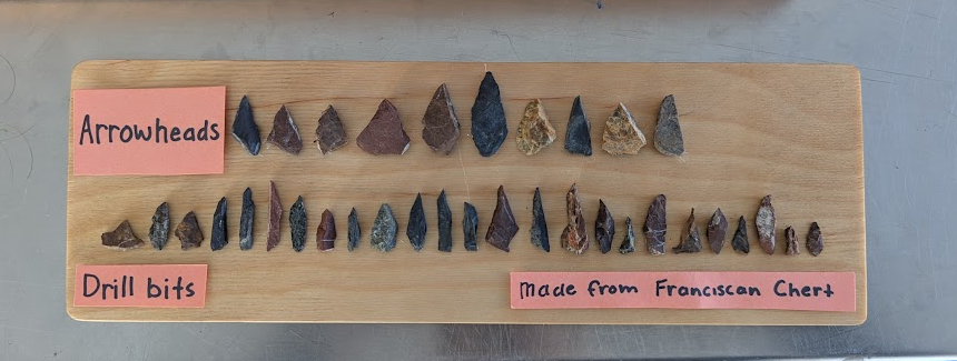 A wooden board displaying rows of arrowheads and drill bits made from Franciscan chert