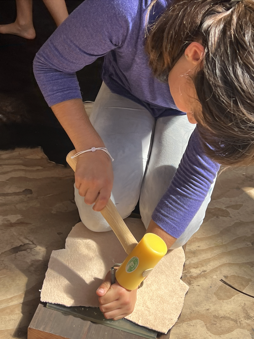 A child using a yellow mallet to work leather on a wooden board