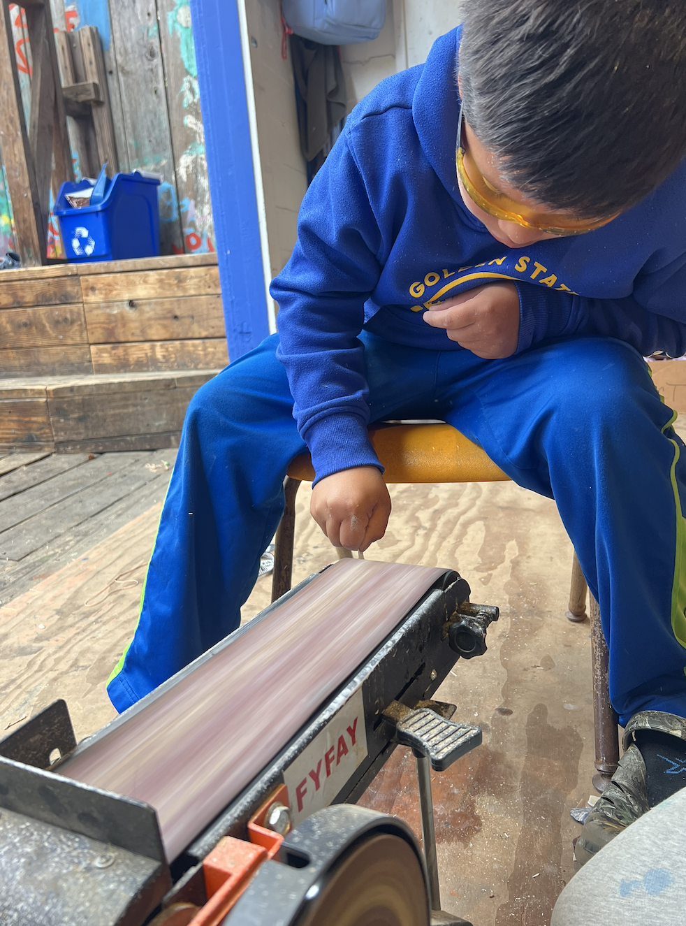 A child wearing safety glasses using a belt sander to shape a piece of stone into an arrowhead