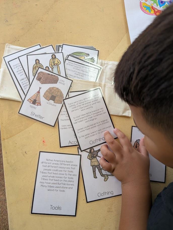 A 2nd grader reading information cards about Native American shelter, clothing, and tools spread across a table